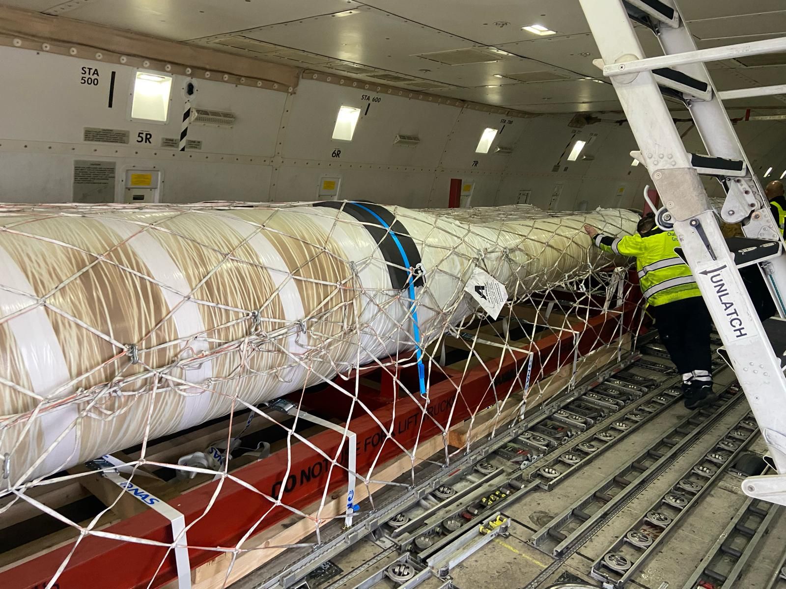 A long wrapped tubing bundle strapped to a red pallet inside the main deck of a cargo aircraft, with a handler in a high-visibility jacket alongside.