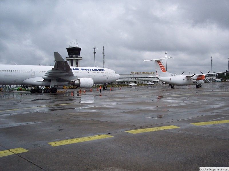 Two widebody aircraft parked on a wet apron at Port Harcourt International Airport, with the control tower behind and ground crew in high-visibility vests.