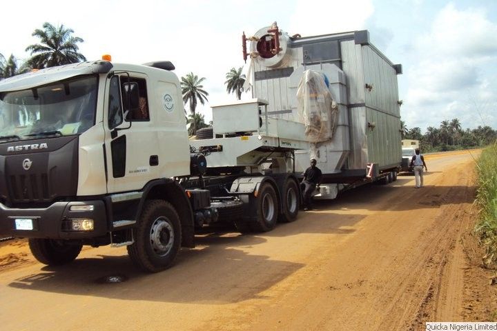 A white tractor unit hauling a large wrapped boiler on a low trailer along an unsurfaced laterite road lined with palms.